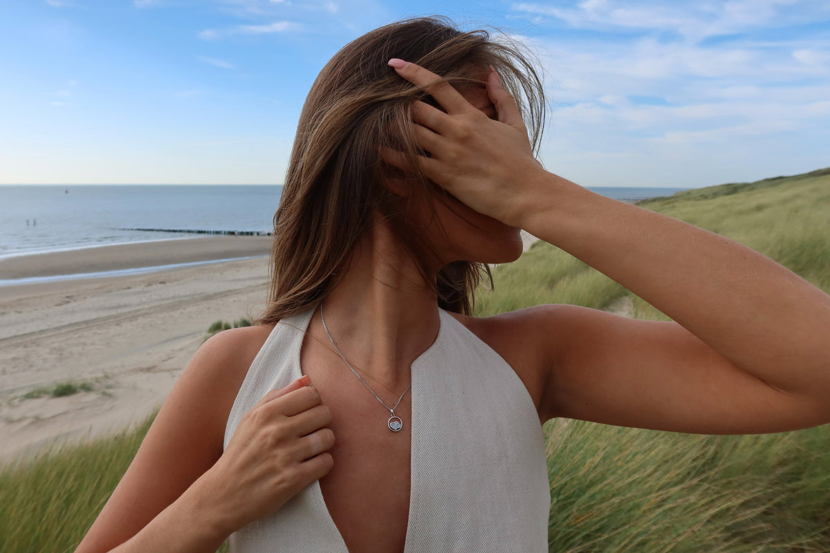 Hanger van massief witgoud met een ronde moedermelkhars en een subtiele gravure, gedragen door een vrouw op het strand.