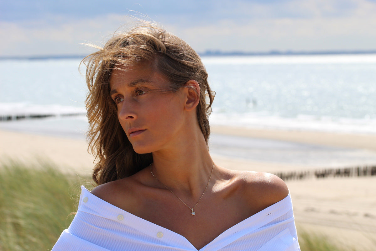 Hanger van 925 zilver met een klein, transparant stuk moedermelkhars, gedragen door een vrouw met een off-shoulder blouse, op een strandachtergrond.