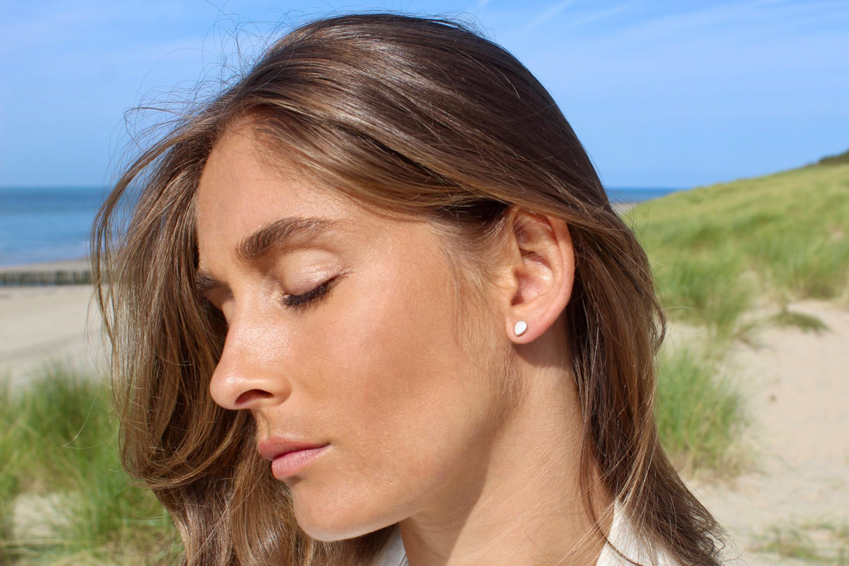 Massief gouden oorstekers in een eenvoudige druppelvorm, gedragen door een vrouw met een natuurlijke uitstraling, tegen een strandachtergrond.