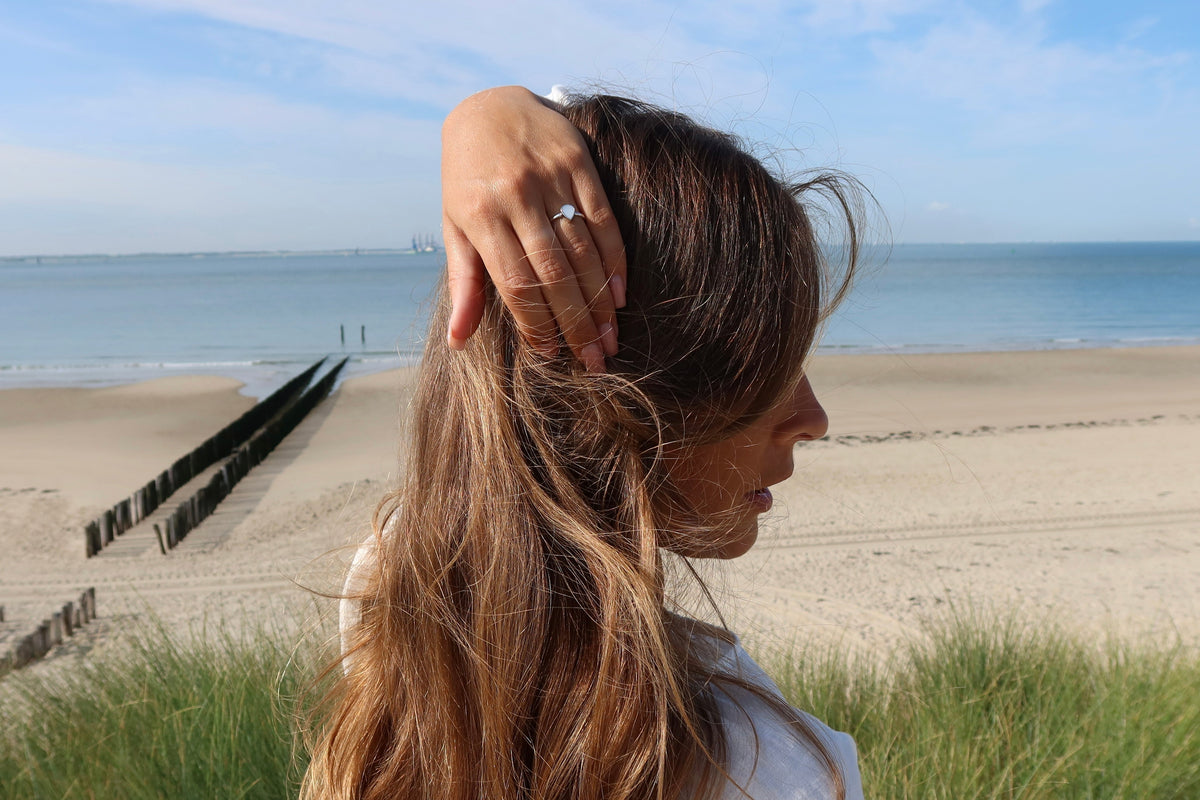 Moedermelk-ring in massief witgoud, met een glanzend oppervlak, gedragen door een vrouw met lang bruin haar, op een strandachtergrond.