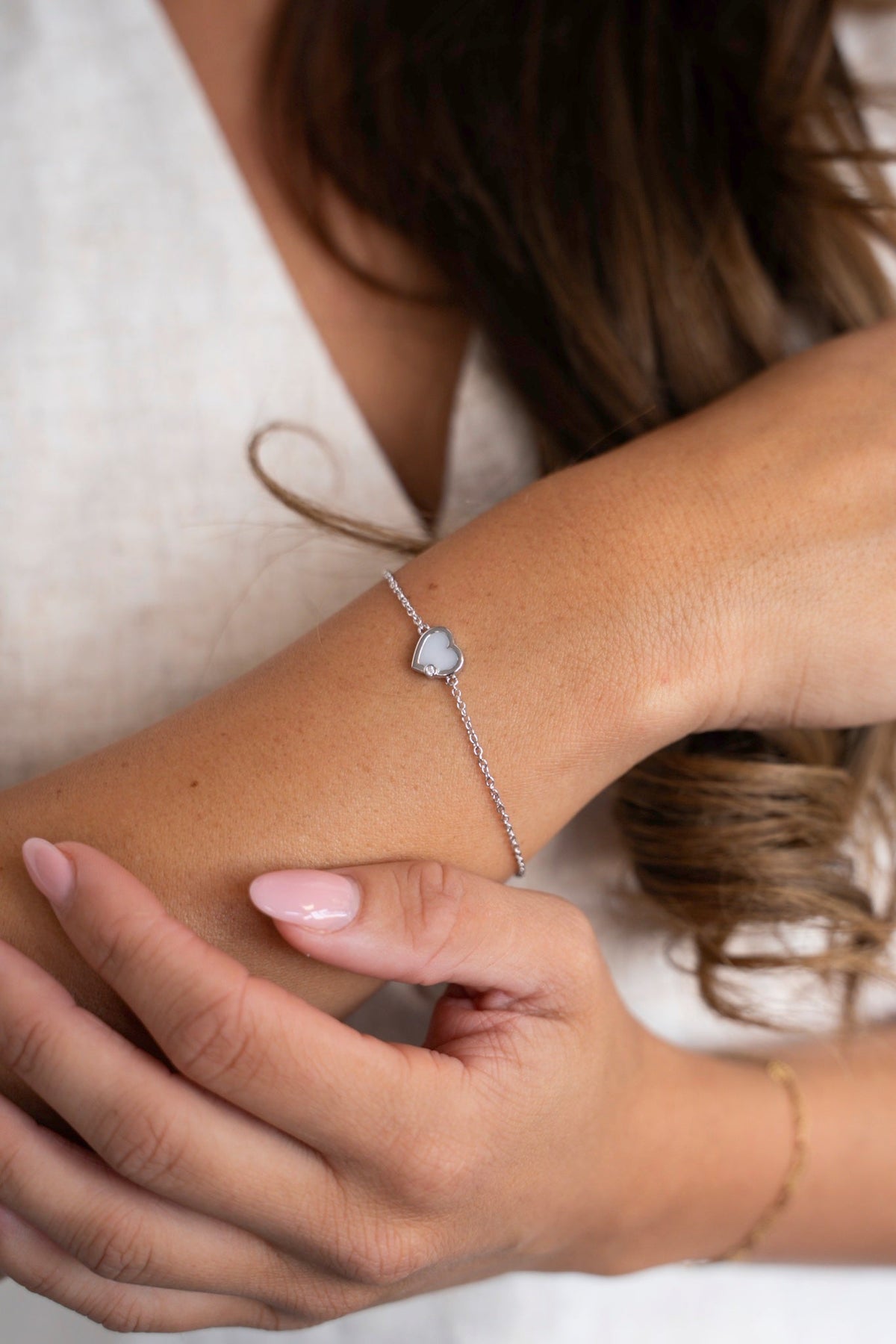 Esmée-armband van massief witgoud met moedermelkhartje, gedragen om de pols tegen een linnen top.