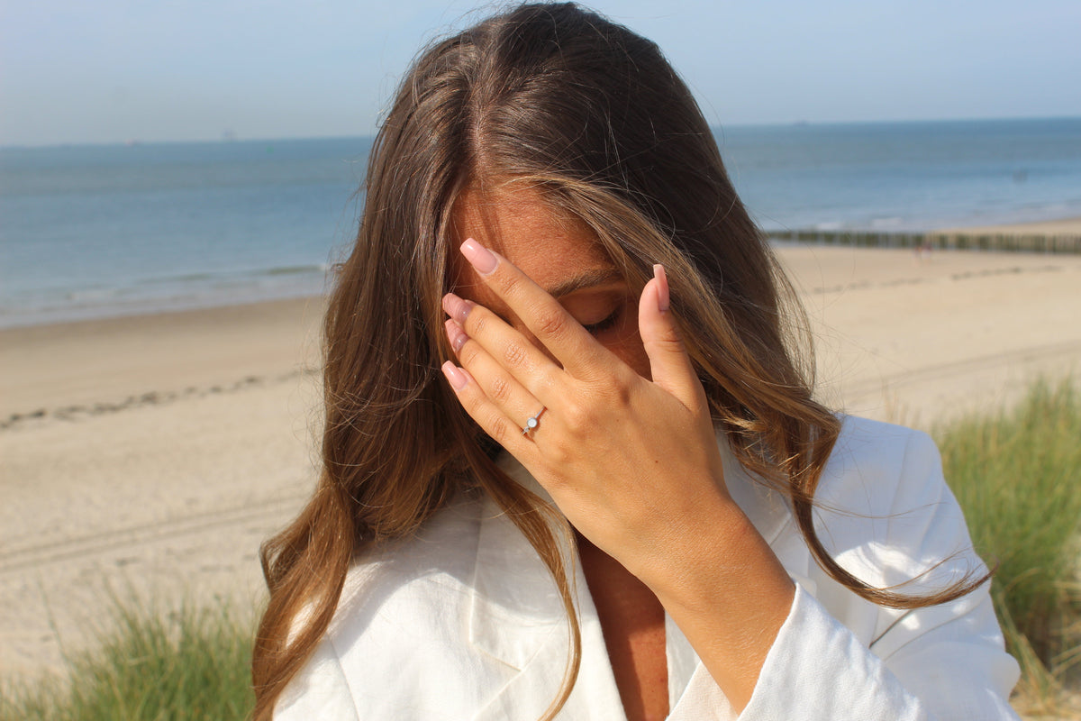 Massief gouden ring met een heldere steen van moedermelkhars, gedragen door een vrouw met golvend haar, op een strandachtergrond.