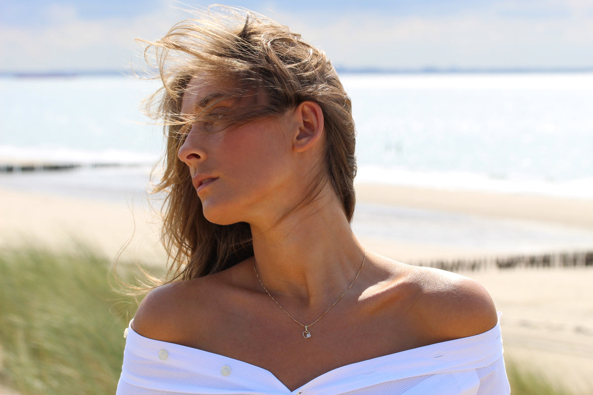 Hanger in massief roségoud met een hartvormige moedermelkhars, gedragen door een vrouw met een off-shoulder blouse, tegen een strandachtergrond.