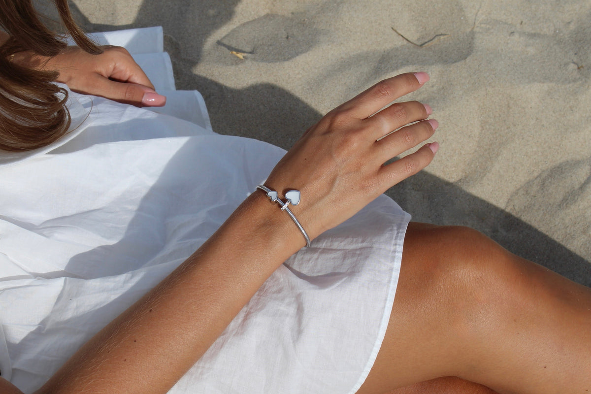 Zilveren bedel in de vorm van een hart, gemaakt van moedermelkhars, bevestigd aan een armband, gedragen door een persoon met een witte blouse.