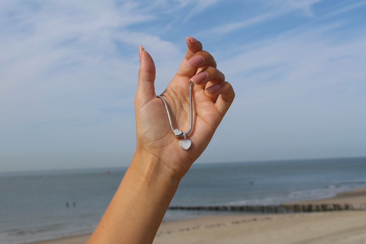 Hangertje in massief witgoud met een hartvormige moedermelkhars en een geboortesteen, vastgehouden door een hand tegen een strandachtergrond.
