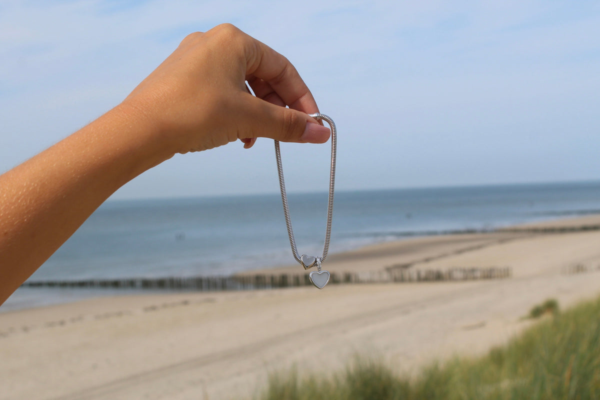 Hangertje in massief witgoud met een hartvormige bedel, vastgehouden door een hand tegen een achtergrond van zee en strand.