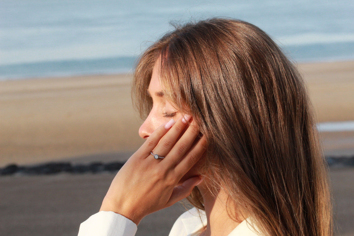 Massief roségouden ring met een vlak van moedermelkhars, gedragen door een vrouw met lang bruin haar, op een strandachtergrond.