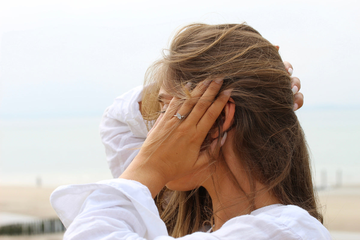 Massief roségouden ring met een ingelegde steen van moedermelk, gedragen door een vrouw met lang haar, op een strandachtergrond.