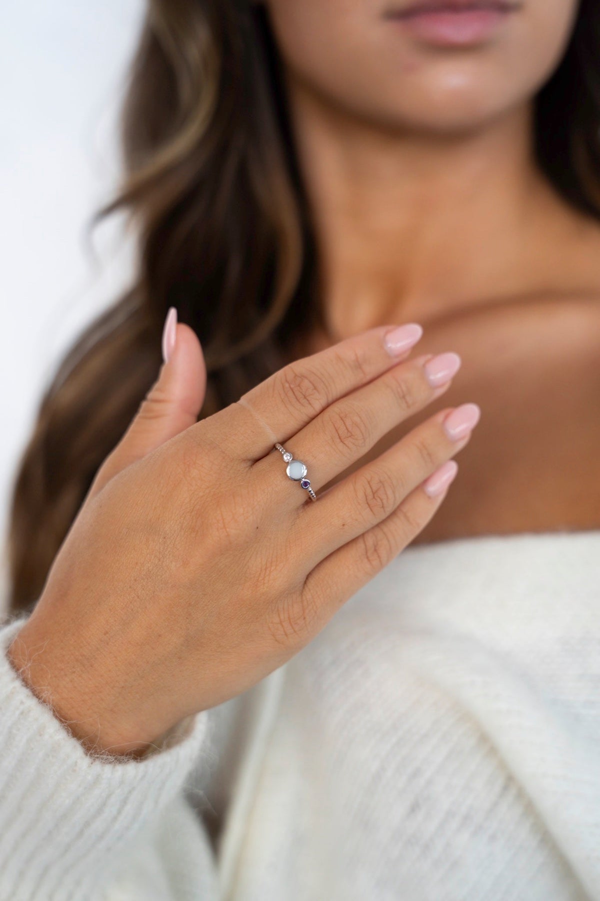 Hand met de witgouden Lot-ring met moedermelk en geboortestenen, tegen een witte gebreide trui.
