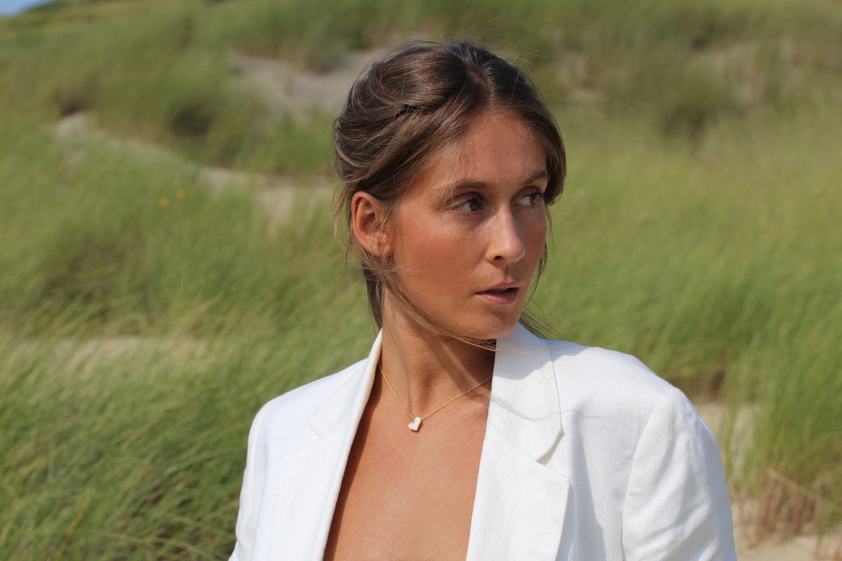 Hanger van massief geelgoud met een subtiele vorm, zichtbaar in de zon, gedragen door een vrouw in een witte blazer.
