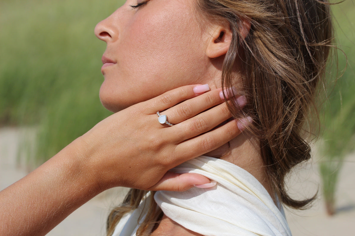 Massief geelgouden moedermelk-ring met een ronde, witte harssteen, gedragen door een vrouw die haar hals aanraakt.