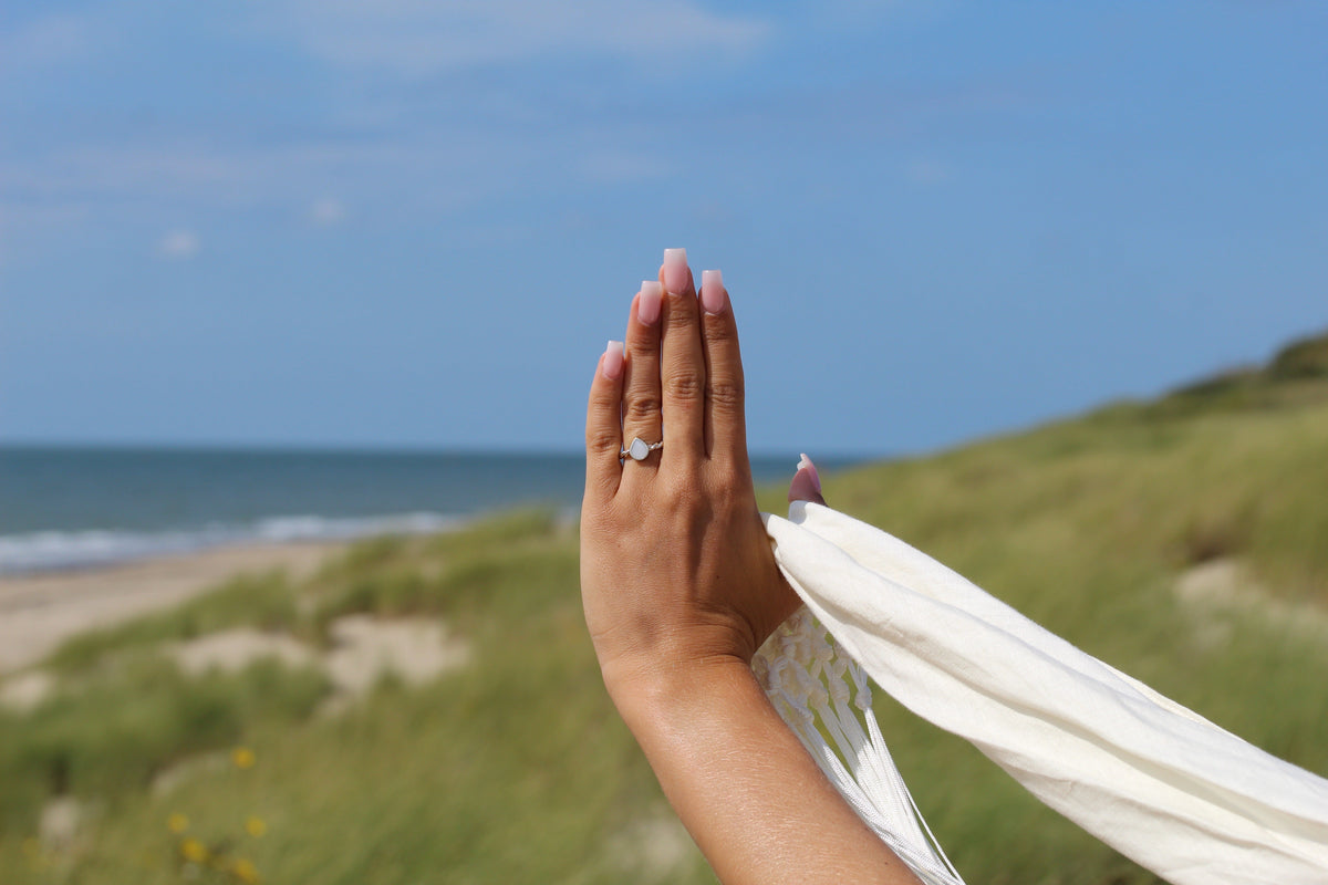 Massief gouden ring met een druppelvormige moedermelkhars en een subtiele glans, gedragen op een hand met lange nagels, tegen een natuurlijke achtergrond van zee en gras.
